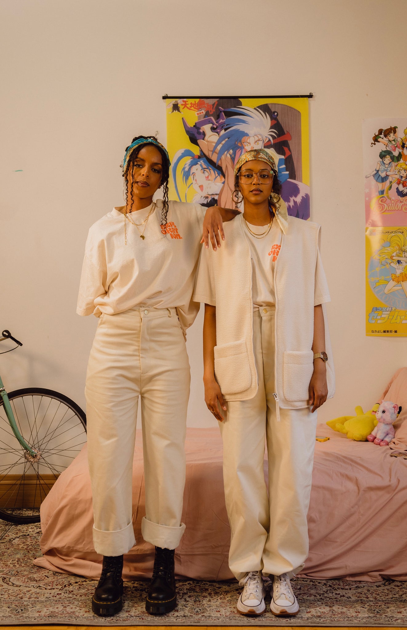 Two girls standing in anime-themed room. Both are wearing cream oversized tee, cream baggy jeans and silk bandana. One girl is wearing oversized cream vest on top of tee.